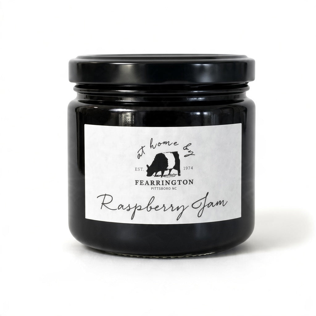 A glass jar of raspberry jam is displayed with a black and white striped ribbon adorning the lid. The white label reads "Fearrington Housemade Raspberry Jam" with a list of ingredients: raspberry, vanilla, lemon, pectin, sugar. The text also indicates it is handmade and may contain allergens. Perfect as a hostess gift!