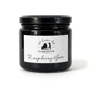 A glass jar of raspberry jam is displayed with a black and white striped ribbon adorning the lid. The white label reads "Fearrington Housemade Raspberry Jam" with a list of ingredients: raspberry, vanilla, lemon, pectin, sugar. The text also indicates it is handmade and may contain allergens. Perfect as a hostess gift!