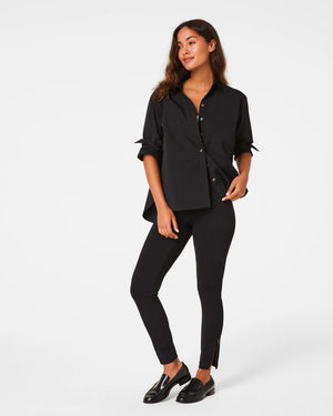 A woman with long wavy hair poses against a plain white background wearing a loose black button-up shirt, SPANX PONTE ZIP LEGGING by SPANX with ankle slits, and black loafers. Her sleeves are rolled up for a relaxed look.