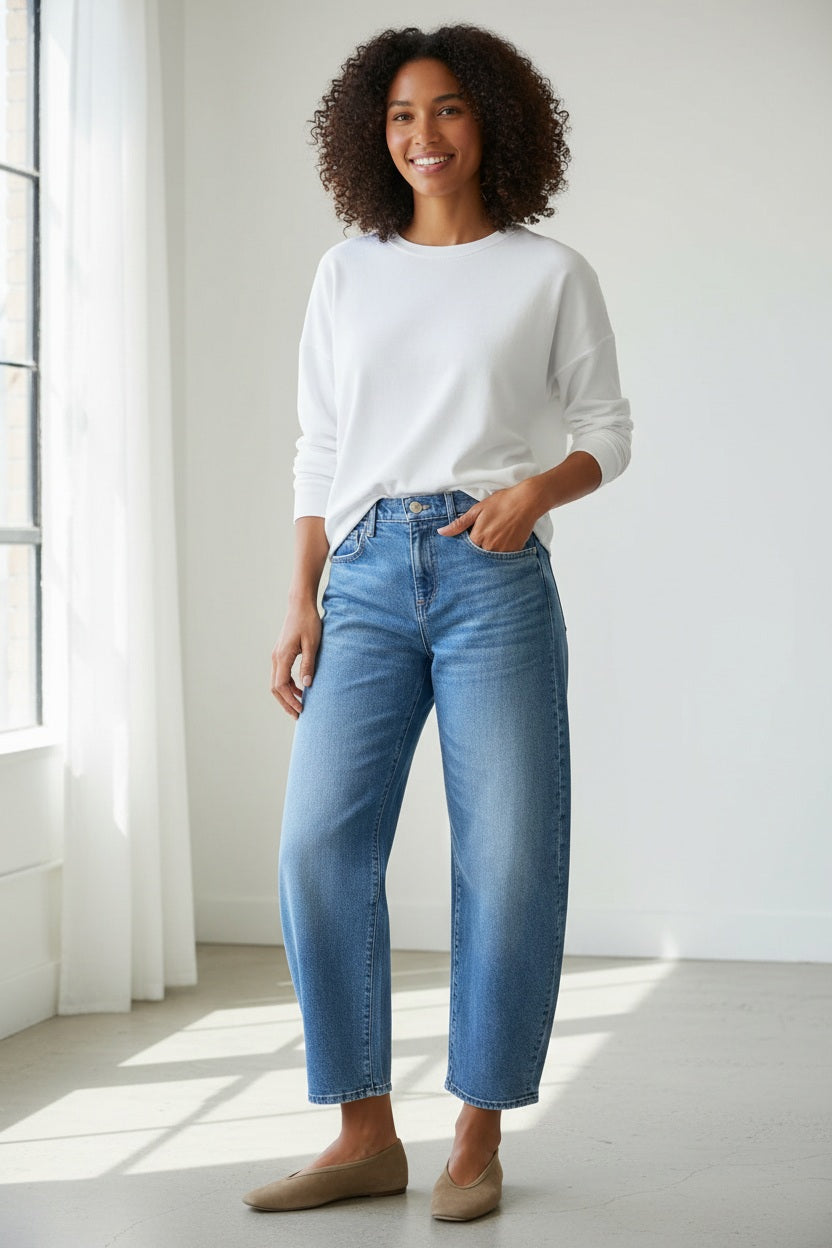 A person stands in a kitchen with their back to the camera, wearing FRANK &amp; EILEEN’s Brennan Everyday Barrel Jean in 1978 Vintage Wash, a white long-sleeve shirt, and brown shoes. The kitchen has light cabinets, a dark oven, and wood floors.