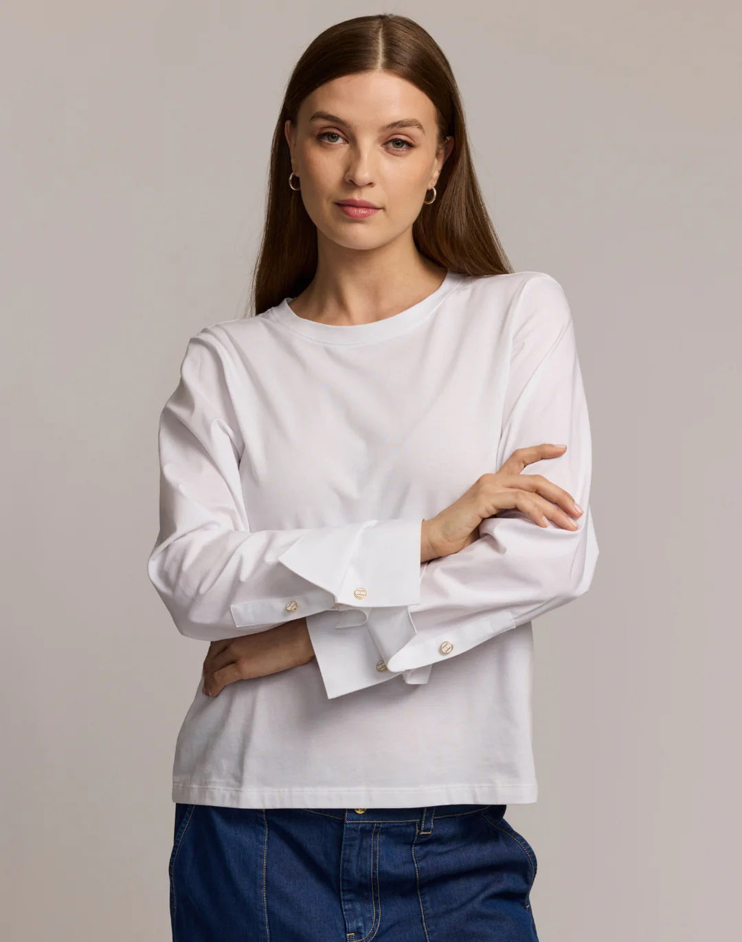 A woman with long brown hair wears the HINSON WU REAGEN LONG SLEEVE FOUNDATION LAYER TOP, featuring wide cuffs and gold buttons, paired with blue jeans. She stands against a plain light background with her arms crossed and a neutral expression.