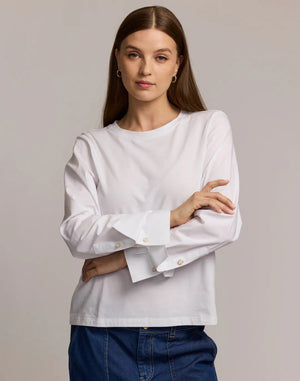 A woman with long brown hair wears the HINSON WU REAGEN LONG SLEEVE FOUNDATION LAYER TOP, featuring wide cuffs and gold buttons, paired with blue jeans. She stands against a plain light background with her arms crossed and a neutral expression.