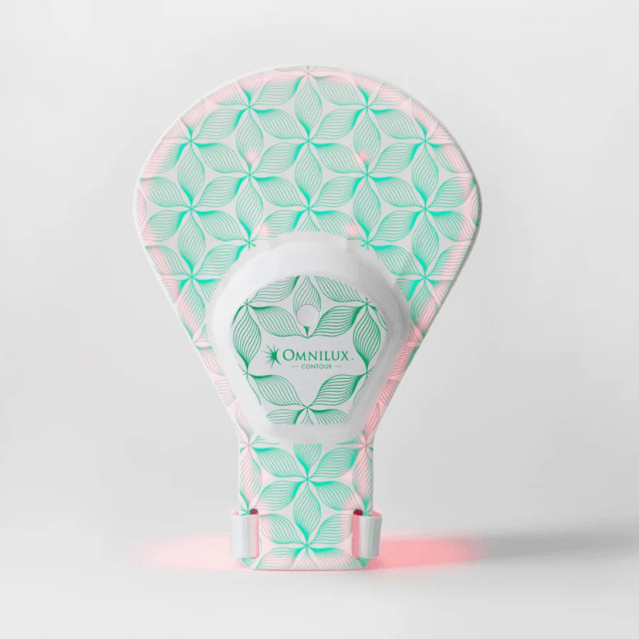 A person wearing a white tank top is using the OMNILUX - CONTOUR GLOVE from OMNILUX, which emits a rejuvenating warm glow to their face. Their expression is calm, standing against a plain background.