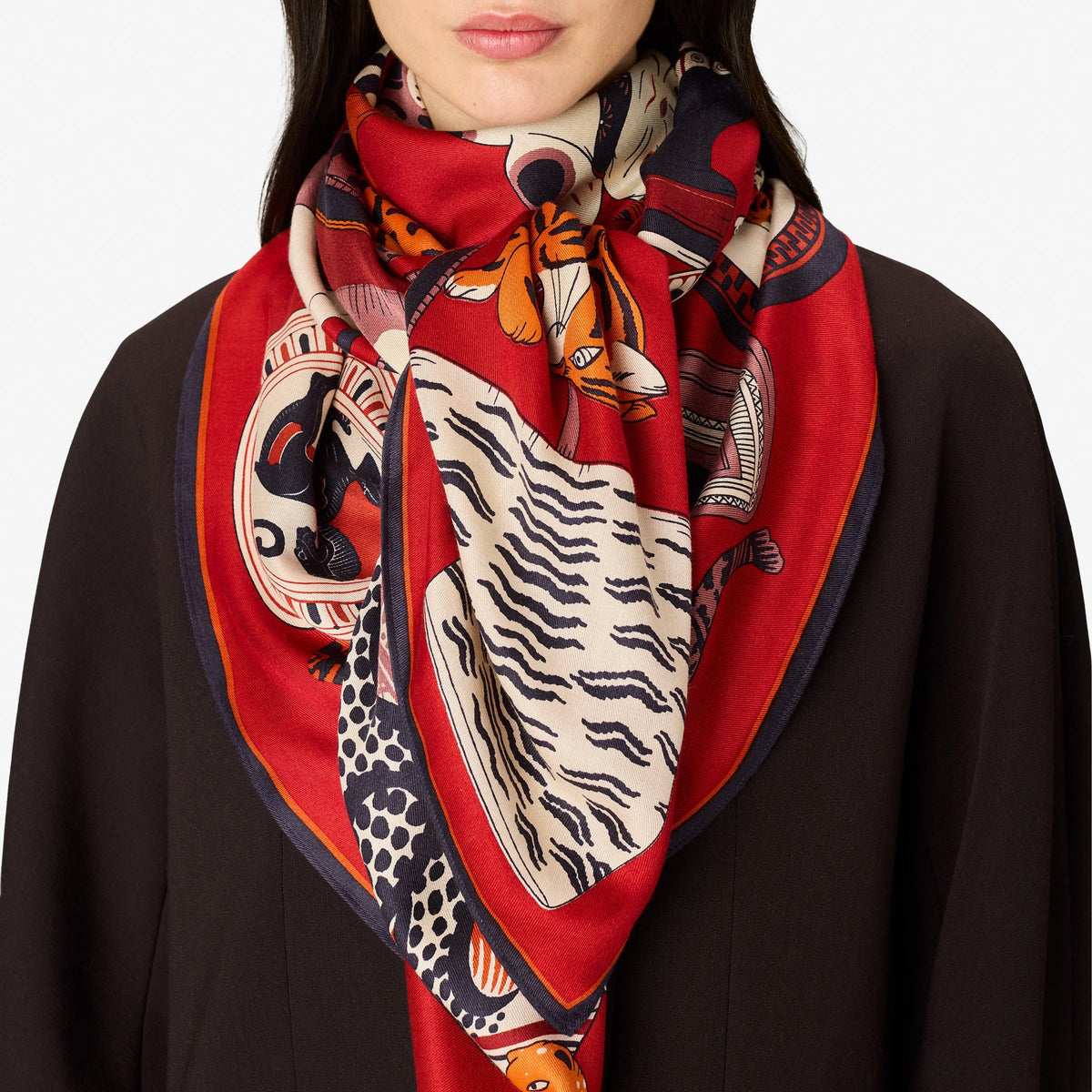 A woman with straight dark hair, dressed in a dark blazer, wears the luxurious INOUI EDITIONS - COLLECTIONNEUR SCARF WITH CATS IN MODAL SILK RED by INOUI. The red scarf—adorned with animal and wave designs and crafted from silk modal— is styled in a triangle around her neck as she stands against a plain white background.