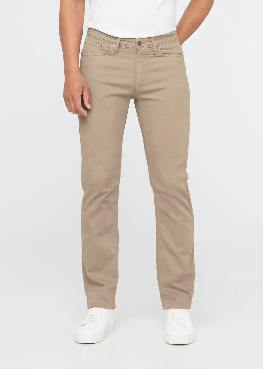 A man with short dark hair stands against a white background, wearing DUER No Sweat Straight Pants in Desert Khaki, a white t-shirt, and white sneakers. He faces the camera with a neutral expression.