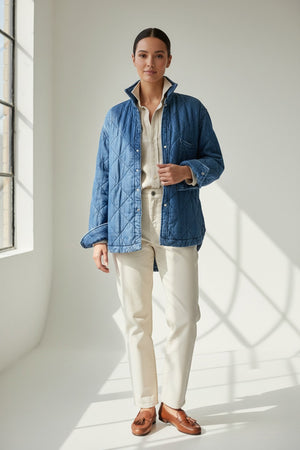 A person holds open a blue FRANK & EILEEN - EILEEN Quilted Shirt Jacket in Famous Denim Distressed Indigo Wash, revealing its interior pocket and distinctive diamond quilted pattern. The person is wearing a cream-colored, ribbed top, with a blurred background.