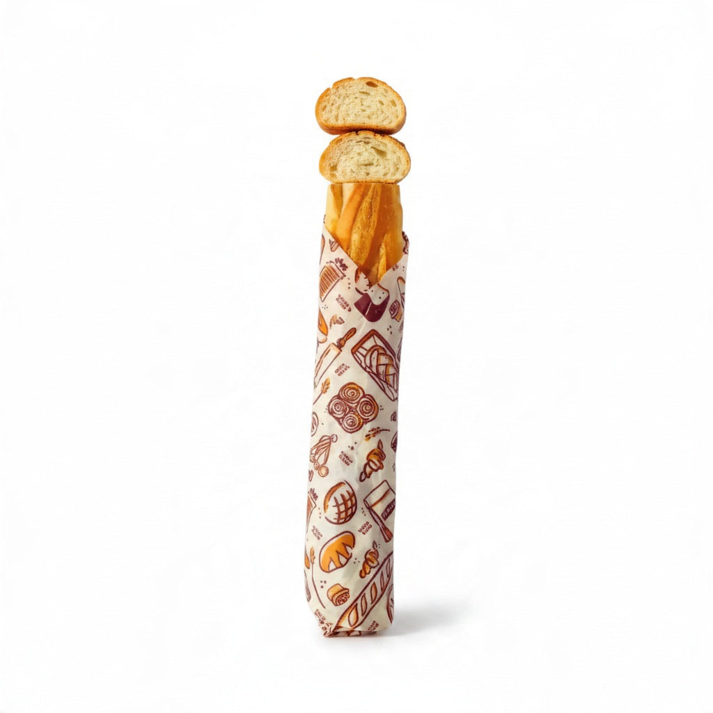 A loaf of bread is partially tucked inside the BEES WRAP- BEESWAX REUSABLE BREAD BAKERY LANE bag by BEES WRAP, featuring a small white label and displayed against a plain white background—a perfect choice for a plastic-free kitchen.
