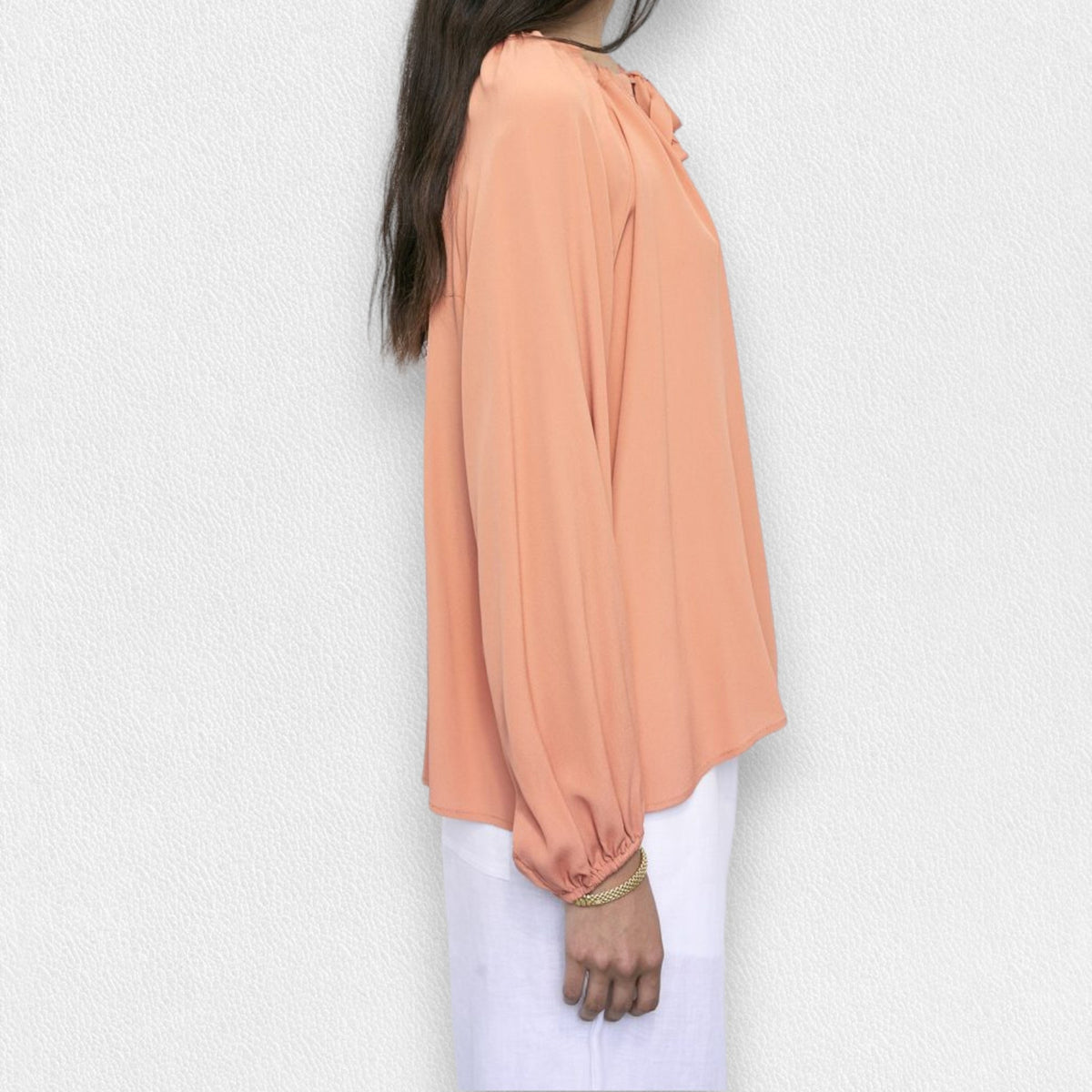 A person wearing a MEIMEIJ - TIE NECK BLOUSE in salmon color, paired with white pants, stands against a light gray textured background. The blouse features long sleeves and a bow at the neckline. The person's face is not visible in the image.