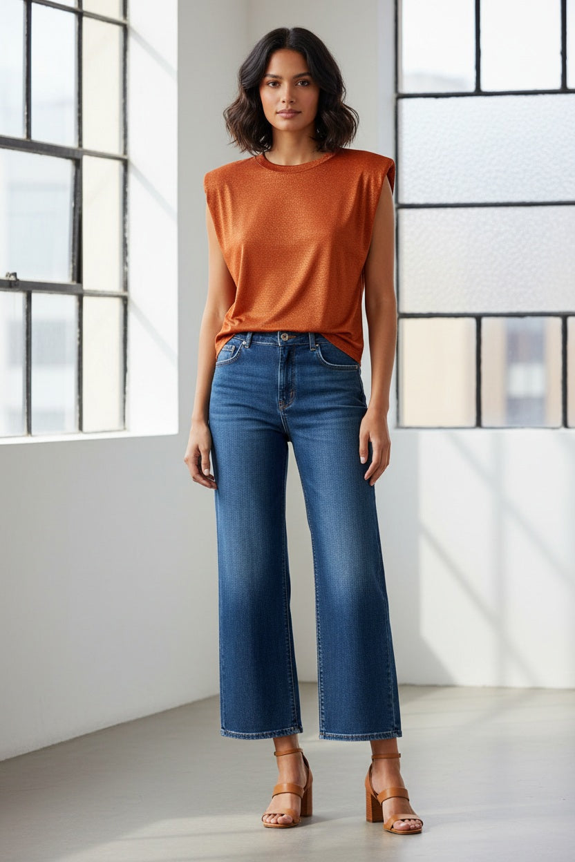 A woman with long brown hair wears the MOMONI LUREX JERSEY SLEEVELESS TOP in green, paired with dark jeans and a tassel necklace, standing against a neutral background.
