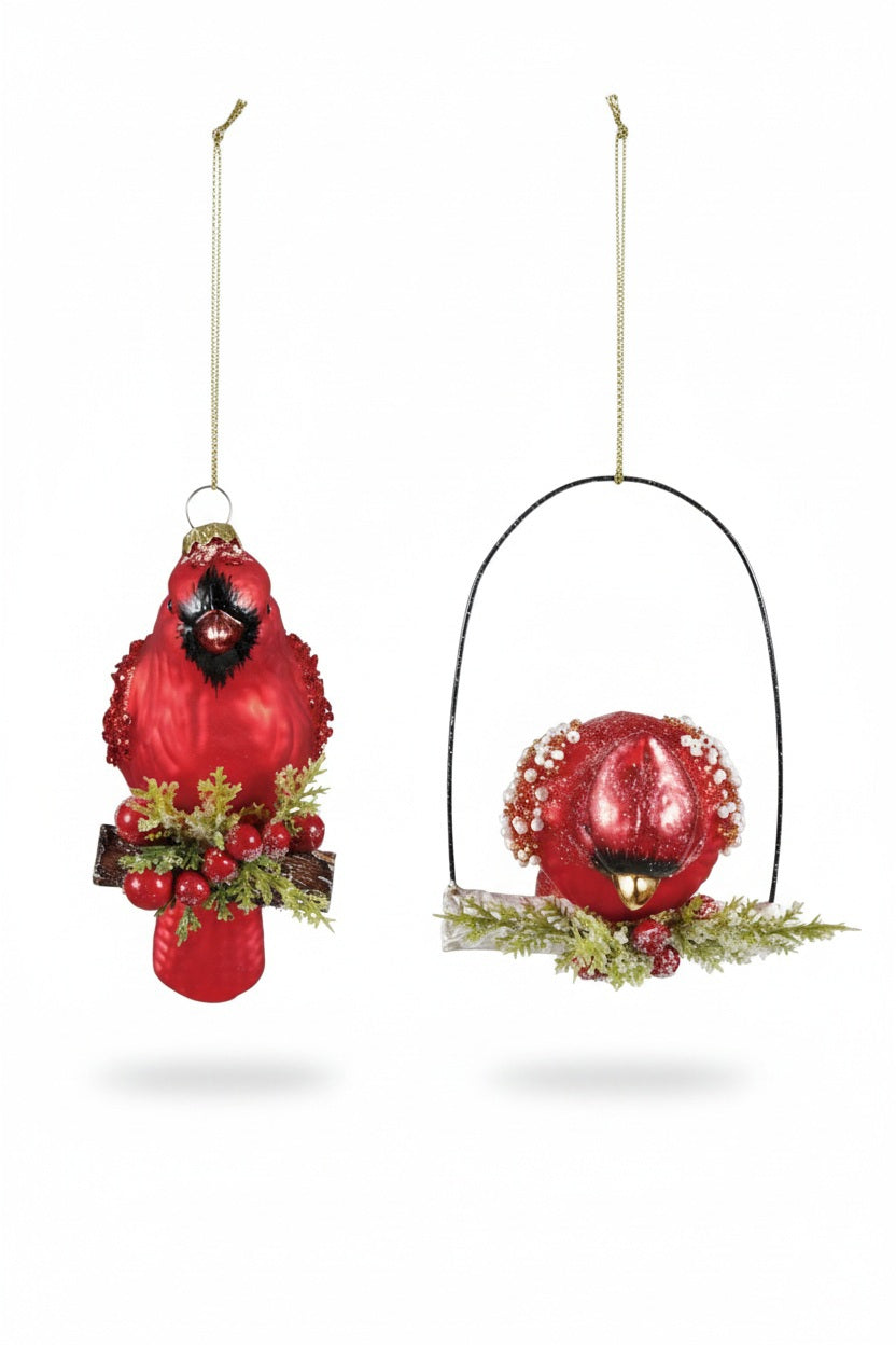 Two Creative Co-op HAND PAINTED CARDINAL ORNAMENT WITH BEADS AND GLITTER pieces hang from a branch, one on a perch and one suspended vertically, both decorated with beads and berries.