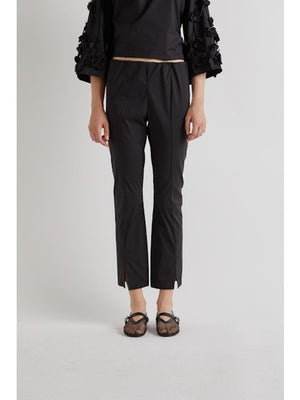 A person stands against a plain background wearing MEIMEIJ FROTN HEM SPLIT COTTON PANT—black, cropped, ankle-length pants with small slits. They also wear a black textured-sleeve top and black shoes. Their face is not visible in the image.