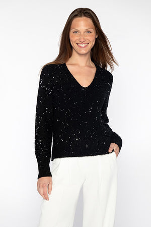 A person wearing the KINROSS CASHMERE - SEQUIN VEE NECK PULLOVER, crafted from sequin yarn, with a V-neck design, paired with white pants stands against a plain white background, smiling with one hand in their pocket.