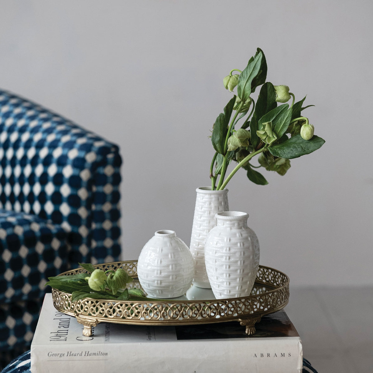 A gold tray with three white vases, including the CREATIVE COOP EMBOSSED STONEWARE BASKET WEAVE VASE, LARGE—two holding green stems—rests on books, with a blue and white patterned chair visible in the background.