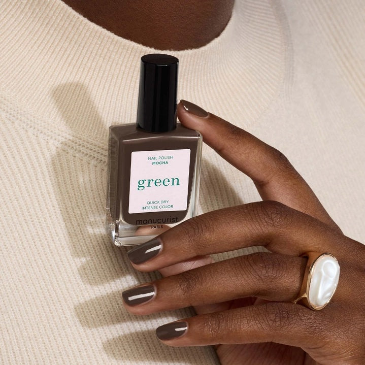 A hand with short nails painted in MANUCURIST - GREEN™ Nail Polish in Mocha by MANUCURIST INC rests on a shiny, metallic, wavy-edged object against a deep red background.
