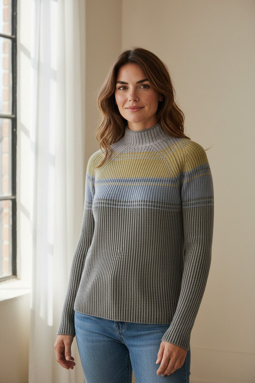A woman with long brown hair, wearing the KINROSS CASHMERE - Plaited Rib Colorblock Funner Cashmere Sweater in Highland Multi by KINROSS, stands by a window in a softly lit room, smiling slightly at the camera.