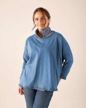 A woman stands barefoot indoors near a large window, looking at the camera with a relaxed expression. She wears the MERSEA - Catalina Ruffle Tee in Steel Blue by MERSEA, featuring delicate feminine ruffle details, paired with blue skinny jeans. Her blonde hair falls loosely around her shoulders.