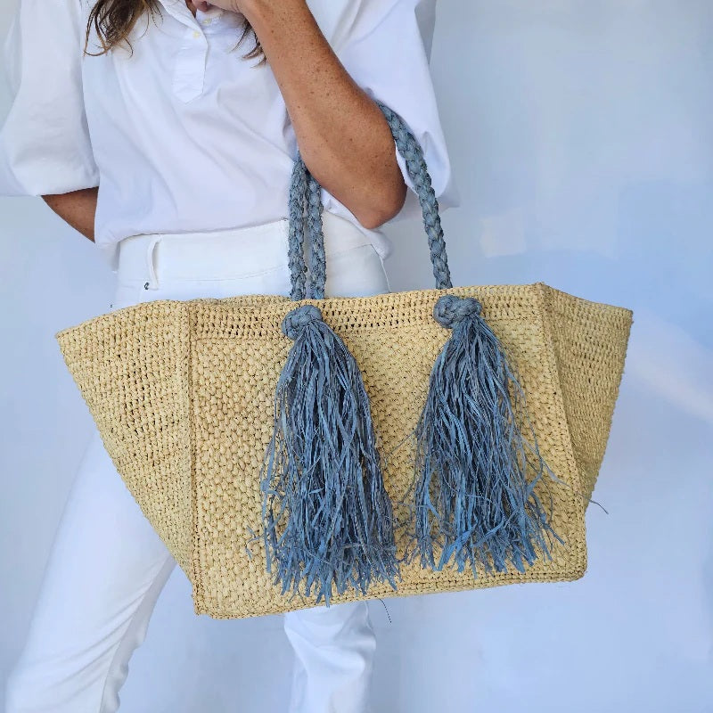 A woman in white shirt and pants holds the BALDIZ Camilla Raffia Tote Bag in Hay and French Blue, featuring chunky braided handles and long blue tassels—a handcrafted accent against a light background.