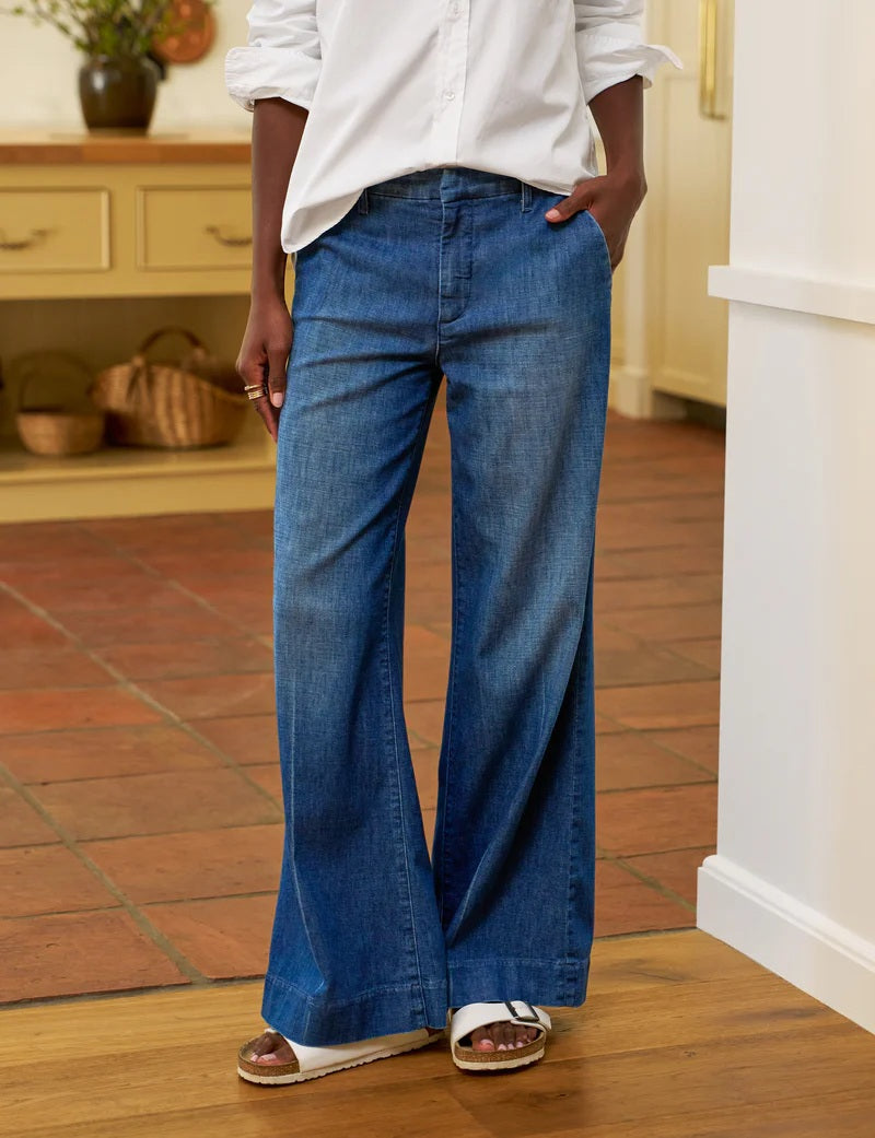 Indoors, someone wears a white long-sleeve shirt, white sandals, and FRANK &amp; EILEEN's WEST VILLAGE NYC WIDE LEG DENIM TROUSER IN 1984 WASH, with their left hand in their pocket. The kitchen background features baskets and wooden cabinets.