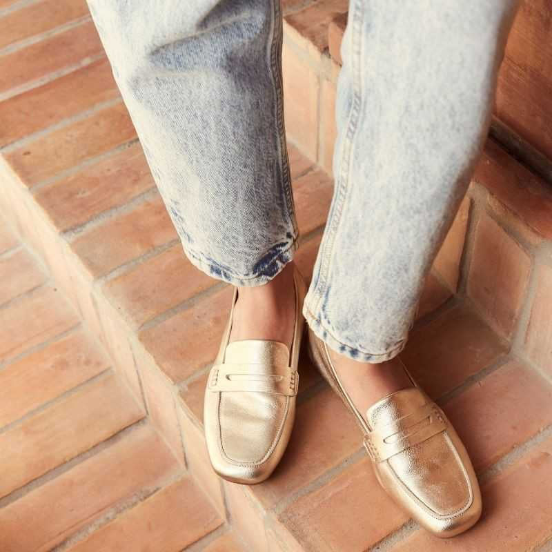 The REQINS - HEMA Metallic Loafer in Platinum features shimmering leather, a low heel, and a decorative front strap, showcased on a plain white background.