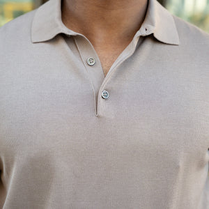 A man in sunglasses wears the COLLARS AND CO MARANELLO Mid-Weight Cotton & Silk 3-Button Polo in Taupe with white trousers, carrying a brown leather bag and phone. Green plants and modern buildings are in the background.