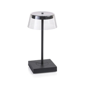 The SARDINIA RECHARGEABLE TABLE LIGHT by ZODAX, with a transparent shade and sleek dark base featuring adjustable brightness, is illuminated on a textured surface beside the book "SHIRO TSUJIMURA: An Art of Living.