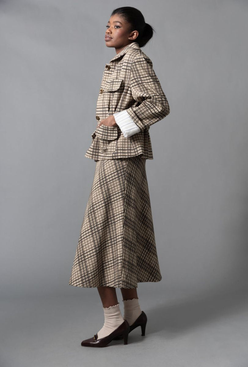 A woman stands sideways against a gray background, wearing the HOPE FOR FLOWERS - FOUR POCKET JACKET by HOPE FOR FLOWERS with a matching beige and brown plaid skirt, white socks, and dark brown heels. Her hair is pulled back as she looks slightly upward.