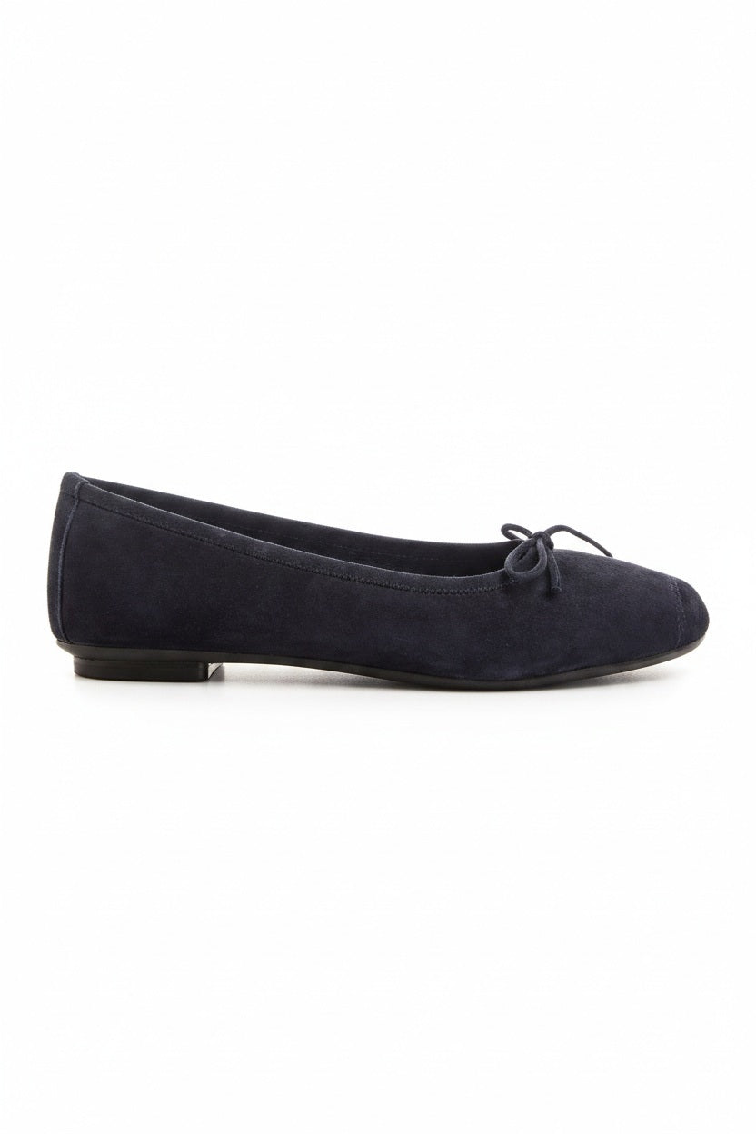 Someone stands on a light floor, one foot slightly raised, wearing REQINS - HARMONY suede ballerina flats in marine with a small bow, paired with blue jeans featuring frayed hems.