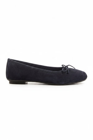 Someone stands on a light floor, one foot slightly raised, wearing REQINS - HARMONY suede ballerina flats in marine with a small bow, paired with blue jeans featuring frayed hems.