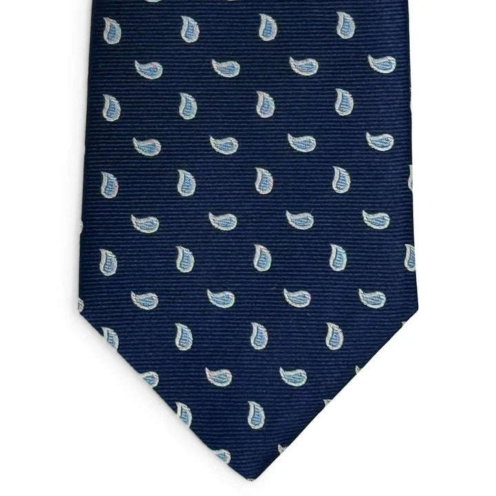 A rolled-up PETER-BLAIR - CAPE COD SILK TIE IN NAVY AND BLUE by BIRD DOG BAY, handmade in New York, featuring a navy blue silk fabric with a repeating light blue and white paisley pattern, displayed on a white background.