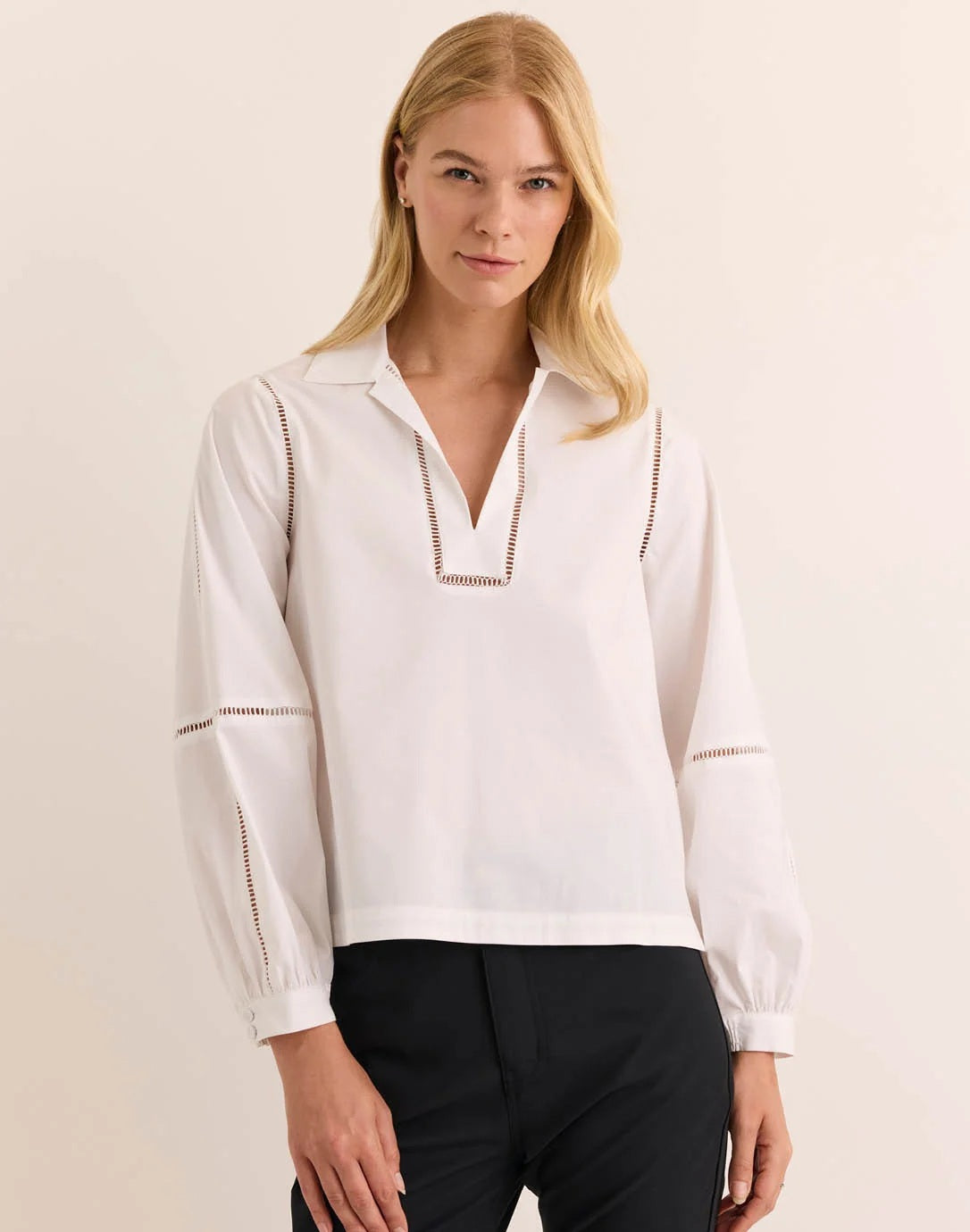 A woman with long blonde hair wears the HINSON WU LAURA Long Sleeve Lace Trim Top in white, crafted from stretch polished cotton and featuring decorative cut-out lace details along the sleeves and neckline. She stands against a neutral background, looking toward the camera with a slight smile. This stylish blouse by HINSON WU is also machine washable, offering effortless care alongside its sophisticated design.
