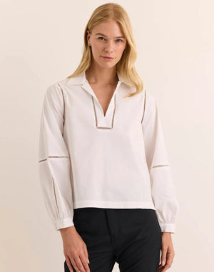 A woman with long blonde hair wears the HINSON WU LAURA Long Sleeve Lace Trim Top in white, crafted from stretch polished cotton and featuring decorative cut-out lace details along the sleeves and neckline. She stands against a neutral background, looking toward the camera with a slight smile. This stylish blouse by HINSON WU is also machine washable, offering effortless care alongside its sophisticated design.
