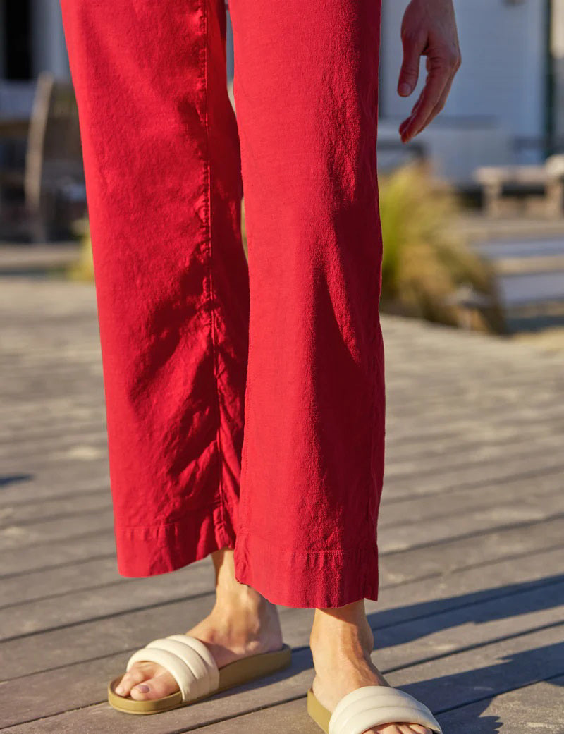 A person stands outdoors on a wooden deck, wearing FRANK &amp; EILEEN’s Wexford Wide Leg Linen Pant in Double Decker Red, a white printed shirt with rolled sleeves, and light-colored slide sandals. The person's face is not visible.