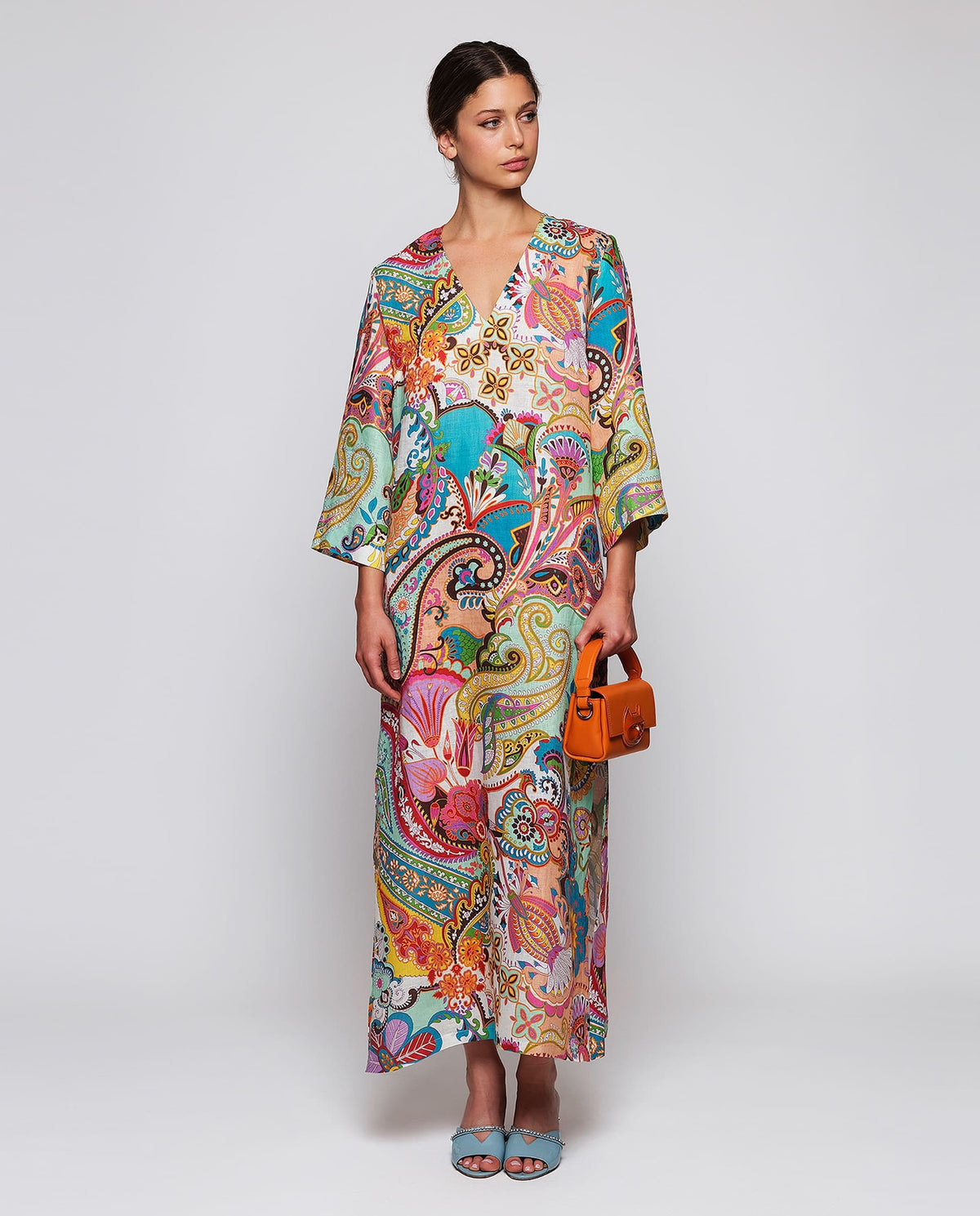 A woman in a MIRTO 1956 Multi Color Paisley Kaftan Style Dress with wide sleeves and V neckline, her hair in a low bun, wears light blue embellished-heel shoes against a plain white background.