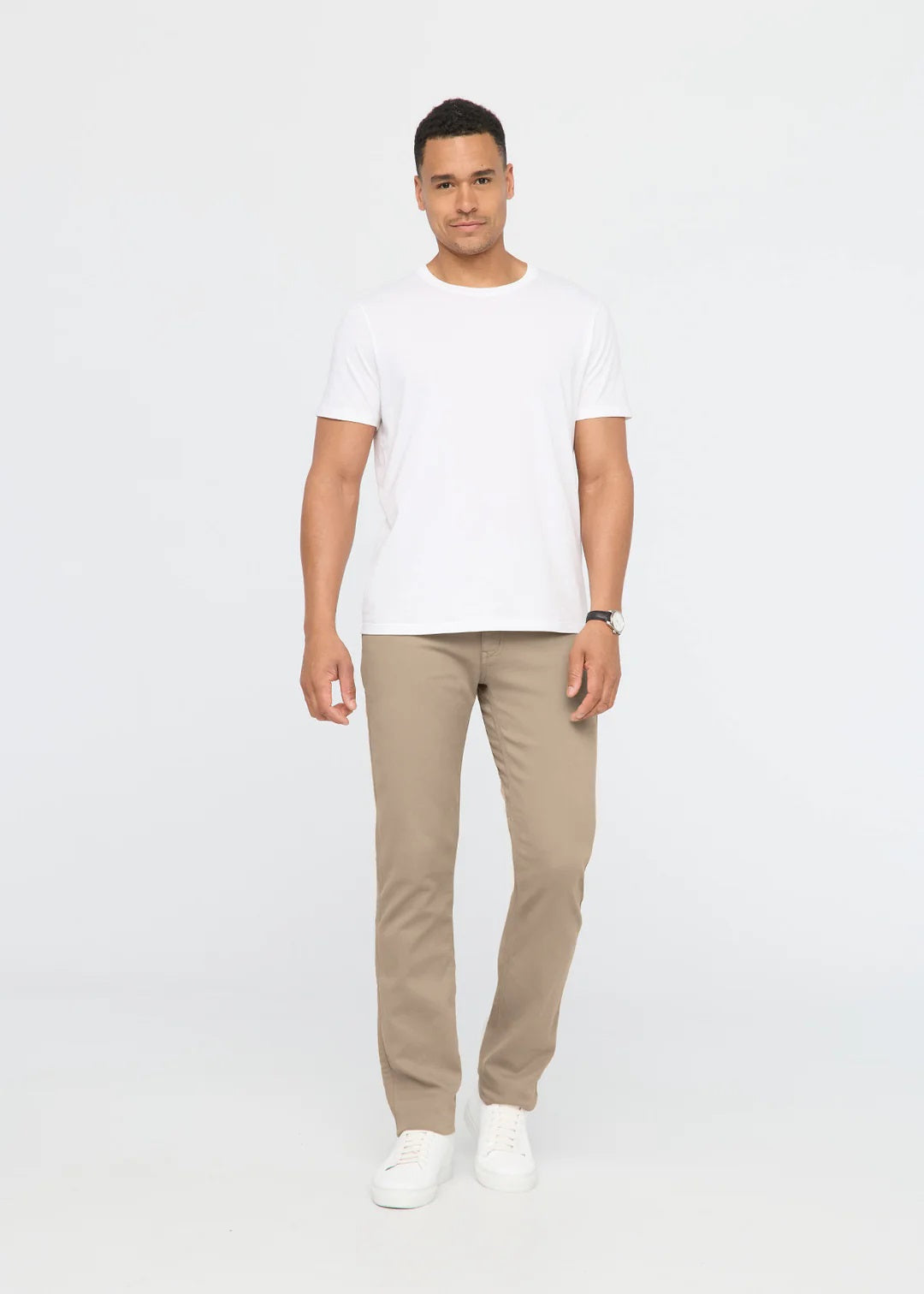 A person wearing DUER NO SWEAT STRAIGHT PANTS IN DESERT KHAKI, a white t-shirt, and white sneakers stands against a plain light gray background.