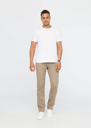 A person wearing DUER NO SWEAT STRAIGHT PANTS IN DESERT KHAKI, a white t-shirt, and white sneakers stands against a plain light gray background.