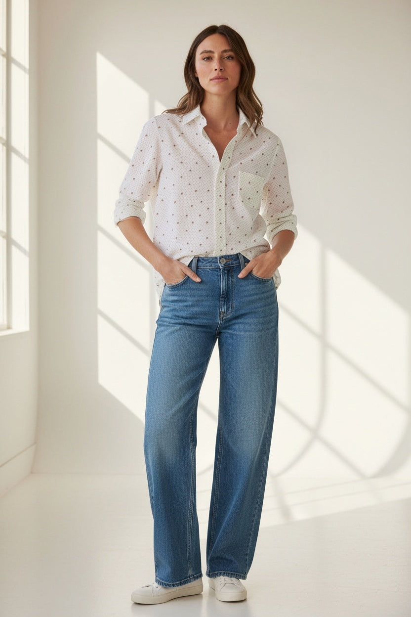 A woman with long wavy hair stands sideways on a porch, looking down, wearing the FRANK &amp; EILEEN EILEEN Relaxed Button Up in Italian Cotton Palm Trees and blue pants. One hand rests in her pocket as the shirt’s relaxed fit drapes effortlessly, enhanced by bust-flattering button placement for subtle shape. The background is bright and green.
