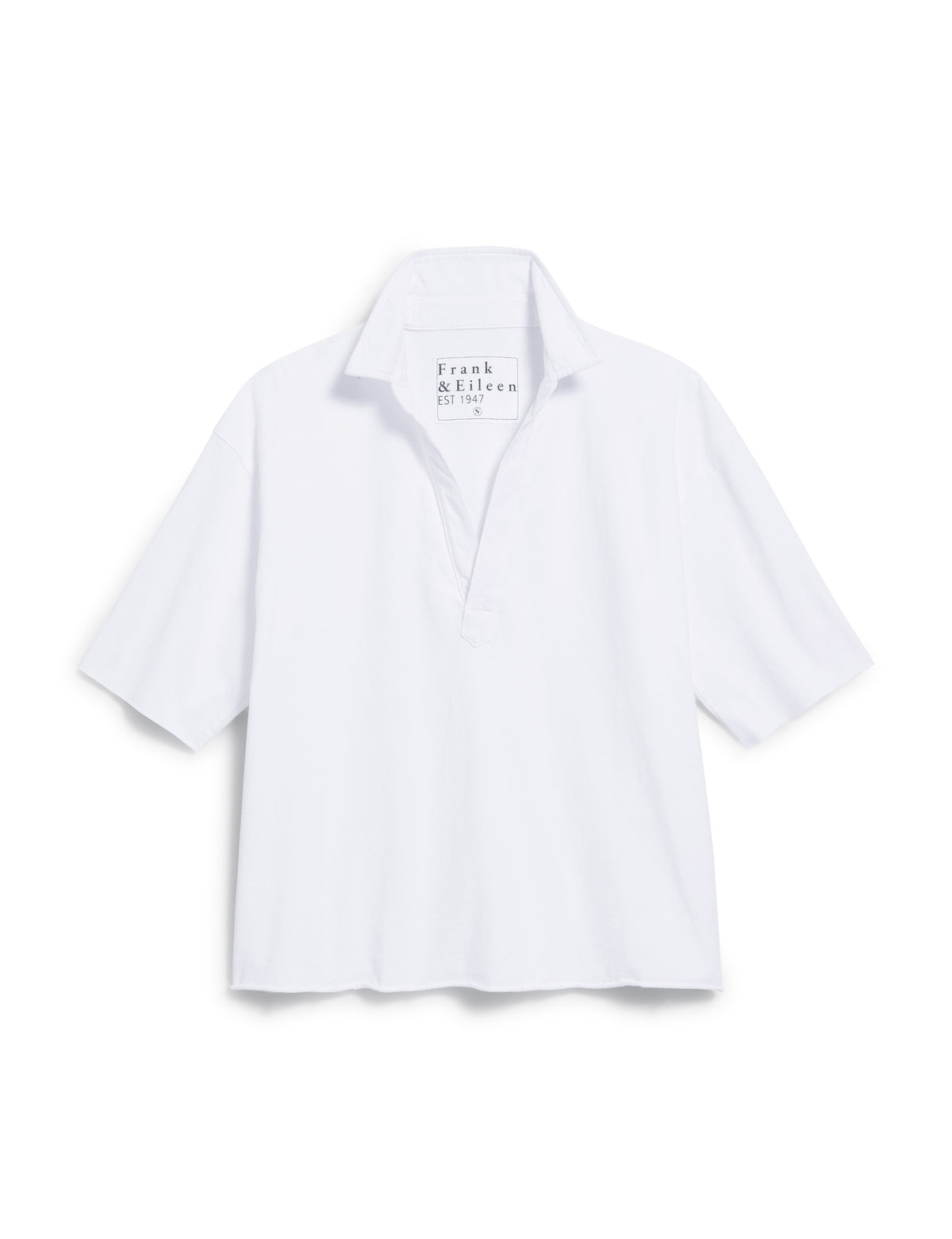A woman in a white blouse and light pants stands on a stone pathway with long brown hair gently cascading as she gazes down gracefully. She wears the FRANK &amp; EILEEN Patty Short Sleeve Popover Henley, perfectly complementing the lush greenery and blurred pool area under soft sunlight.