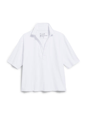 A woman in a white blouse and light pants stands on a stone pathway with long brown hair gently cascading as she gazes down gracefully. She wears the FRANK & EILEEN Patty Short Sleeve Popover Henley, perfectly complementing the lush greenery and blurred pool area under soft sunlight.