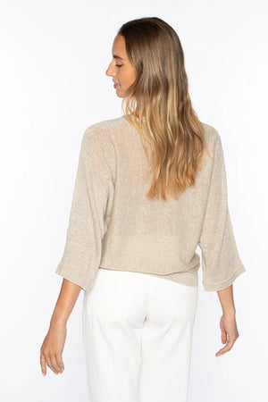 Woman with long, wavy blonde hair models the KINROSS CASHMERE Lurex Dolman Linen Pullover— a loose 100% linen top with three-quarter sleeves—paired with white pants, standing and facing away against a plain white background.