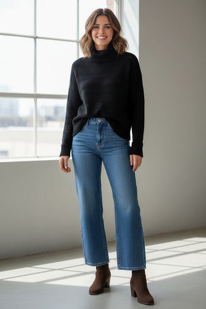 A person with curly hair, dressed in the & ISLA - TEXTURED MOCK NECK PULLOVER IN BLACK by & ISLA and blue jeans, stands against a plain white background facing the camera with a slight smile.