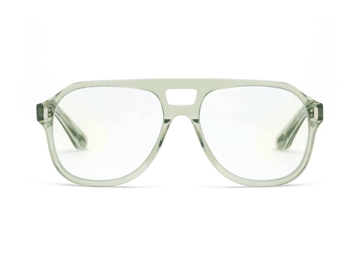 A curly-haired person with glasses from the CADDIS Root Cause Analysis Readers collection sits by a window, holding a mug and smiling. Their aviator-shaped frames catch the light. On the table beside them are a bowl of oranges and a slice of cake. The background shows lush green foliage with a glimpse of the ocean gleaming in the distance.