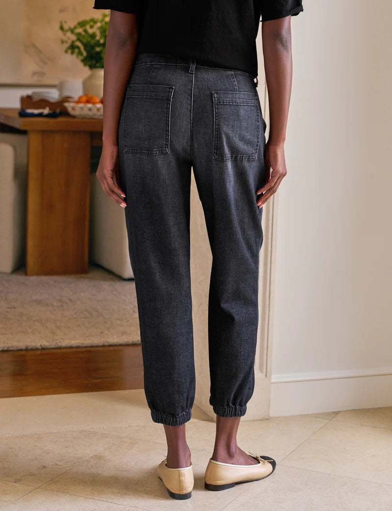 A person stands indoors with hands in the utility pockets of FRANK &amp; EILEEN’s Jameson Jogger Utility Jean in 1981 Wash. They pair it with a black top and tan ballet flats with black toe caps. A dining table and chairs are visible behind them.