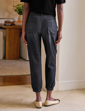 A person stands indoors with hands in the utility pockets of FRANK & EILEEN’s Jameson Jogger Utility Jean in 1981 Wash. They pair it with a black top and tan ballet flats with black toe caps. A dining table and chairs are visible behind them.