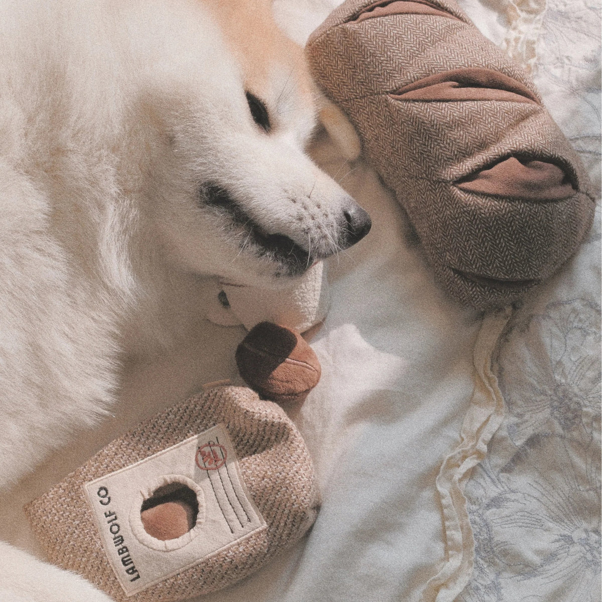 The LAMBWOLF COLLECTIVE - RYE DOG TOY by LAMBWOLF COLLECTIVE is a plush toy shaped like a loaf of bread with three slits on top, cleverly designed as a treat-hiding toy, and shown here resting on a beige and white striped cloth.