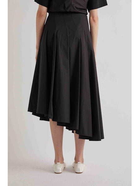 A person is seen from the back wearing a black short-sleeve top, the MEIMEIJ ASYMETRICAL COTTON PLEATED SKIRT, and white flat shoes, against a plain light background.