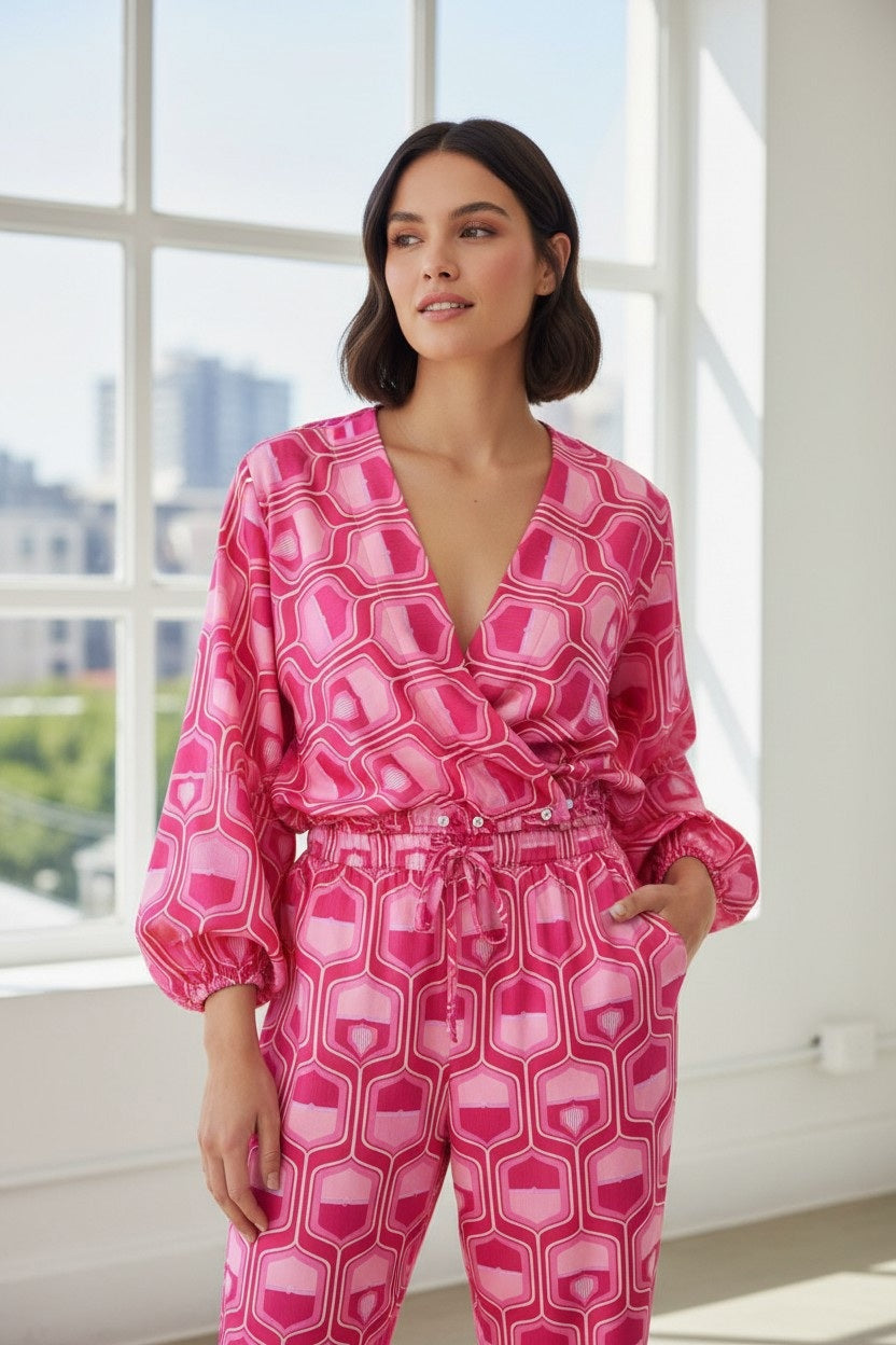 Indoors near large windows with a cityscape in the background, a woman stands looking to the side with her hands in her pockets. She wears the FLOOR - PRINTED BLOUSE by FLOOR, featuring a pink geometric retro pattern, deep V-neck, and long sleeves.