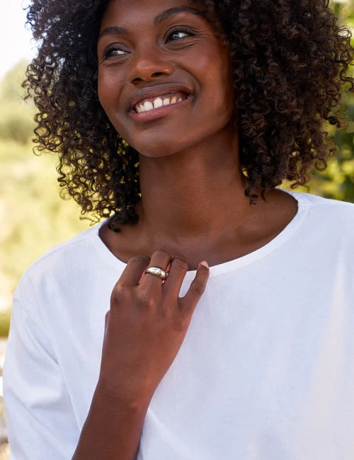 A woman with curly hair smiles outdoors, wearing the FRANK & EILEEN Ella Long Sleeve Boatneck Tee in Heritage Jersey White from the California Heritage Collection, and displaying a ring on her left hand.