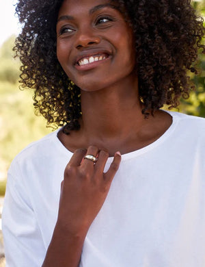A woman with curly hair smiles outdoors, wearing the FRANK & EILEEN Ella Long Sleeve Boatneck Tee in Heritage Jersey White from the California Heritage Collection, and displaying a ring on her left hand.