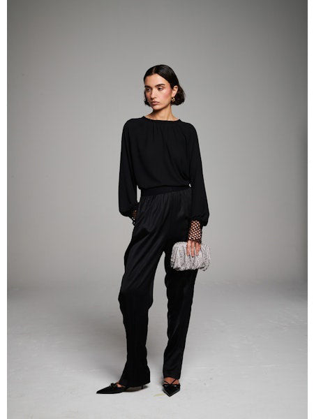 A woman with short dark hair faces away, wearing the MILVA MI - CROCHET CUFF BLOUSE by MILVA, black pants, and pointed black shoes. She holds a silver textured clutch against a plain gray background.