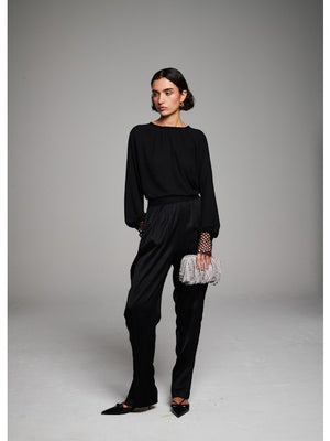 A woman with short dark hair faces away, wearing the MILVA MI - CROCHET CUFF BLOUSE by MILVA, black pants, and pointed black shoes. She holds a silver textured clutch against a plain gray background.